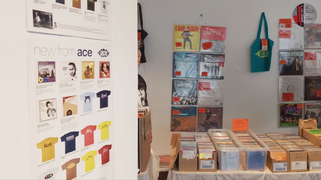Ace Records poster on a wall, and beyond it boxes of CDs on a table, with records and a tote bag hung on the wall behind that table