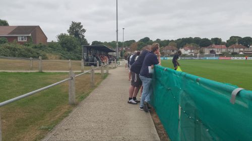 Group of people leaning against hoarding watching football, a football stand in the distance