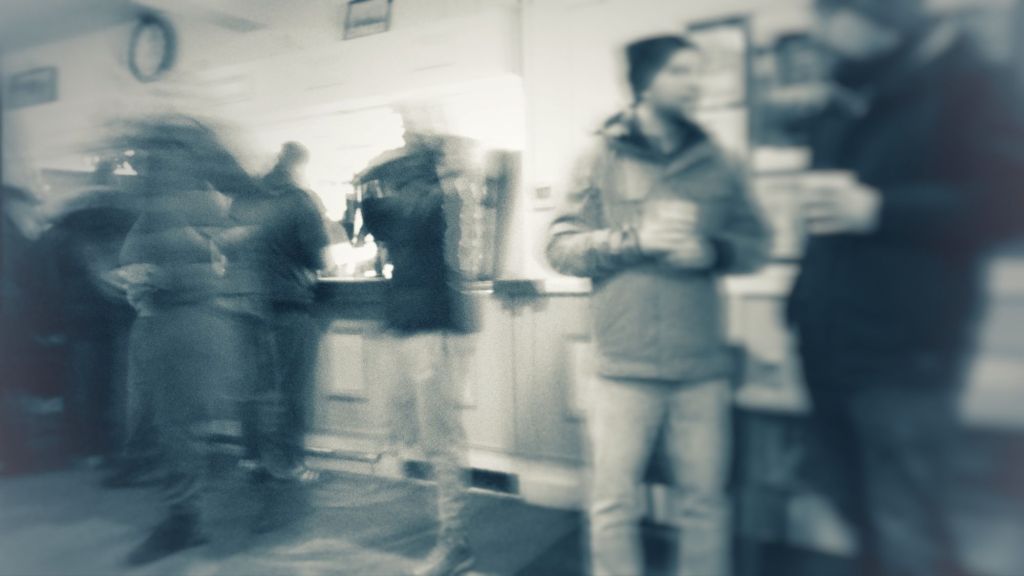 Blurred image of a bar, people are milling about, two men stand by the side with pints of beer