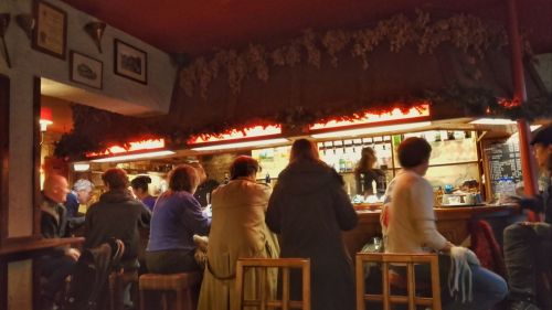 Several people sat at a pub bar, there is a glow behind the bar and a wooden fitting with lighting above. Hops hang from the ceiling.
