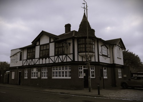 A black and white photo of a pub on the corner of a street. There are some mock tudor features, black bars on white walls. On the corner there is a turret with a pointy roof.