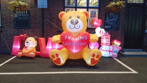 Outside a 1930s style pub there is a massive inflatable Valentine's display. The centrepiece is a huge teddy bear holding a heart that has written on it "Fucking Love You"