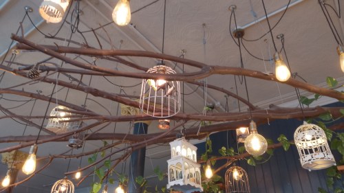 The ceiling of a pub. There is a huge branch reaching across. Various lanterns and light hang from the ceiling and fall between the branches.