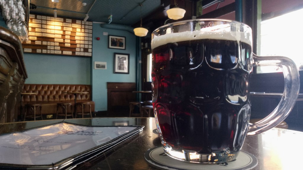 A pint of mild beer in a dimpled jug on a table in a pub. In the distance is the bar, some banquette seating and on the wall a list of available beers.