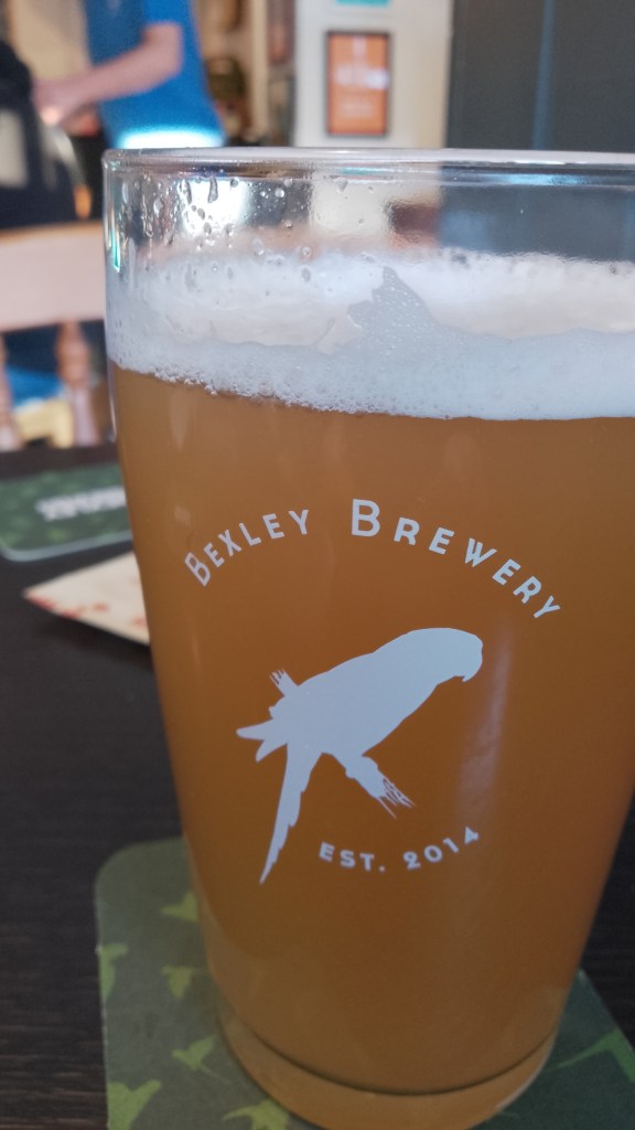 A pint of beer on a beermat on a wooden table. The glass says "Bexley Brewery Est. 2014" and has a silhouette of a parakeet