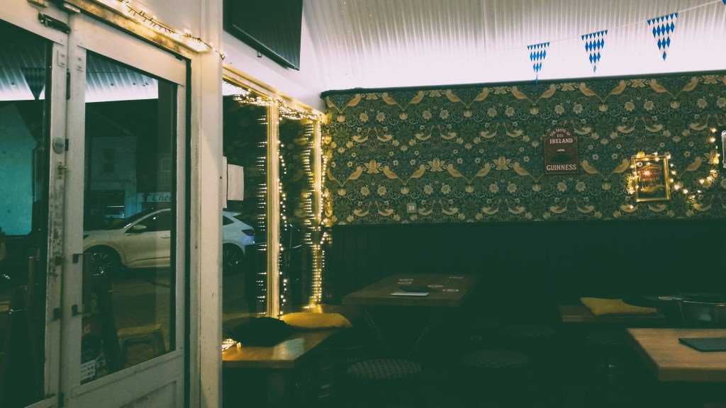Entrance to a craft beer bar. There are wooden tables with yellow cushions. There is green and yellow ornate wallpaper. Blue and white flags hang from the ceiling.