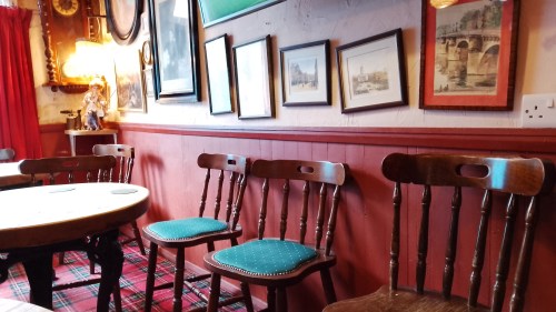 A row of empty chairs in a pub, lined up against a wall. There are old pictures of landscapes on the wall. In the distance is a strange doll with a little stuffed bear