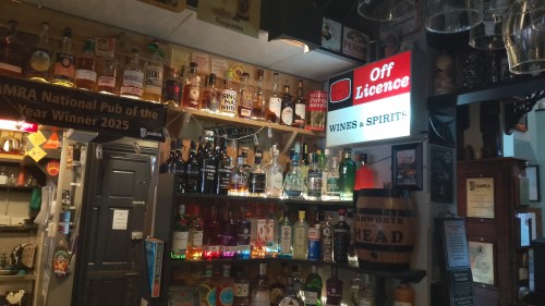 The backbar of the Tamworth Tap. There are a wide variety of spirits on the shelves and retro illuminated sign saying "Off Licence - WINES & SPIRITS"