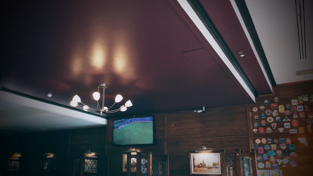 A corner of a Wetherspoons pub. A TV showing football is mounted high up on the wall. There is bright white light fitting hanging from the ceiling. Pump clips are on the wall.